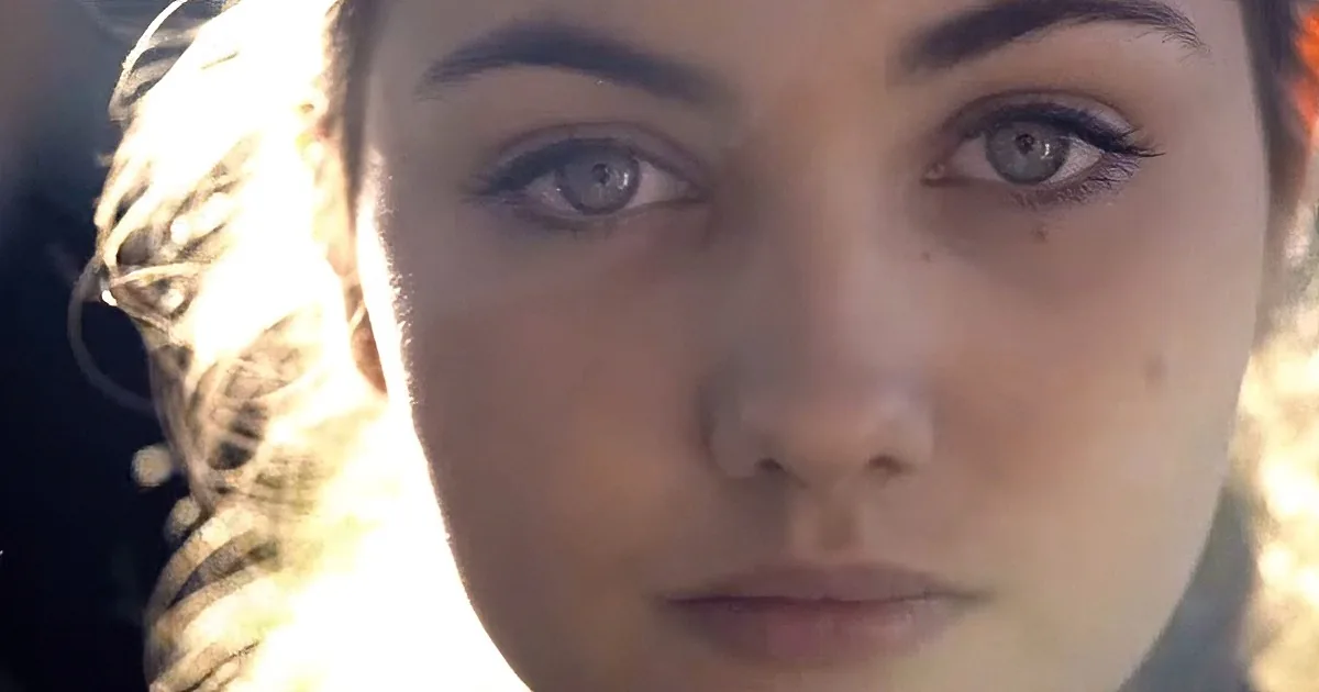 A close-up of a person with light skin, blue eyes, and a nose ring, with sunlight in the background.