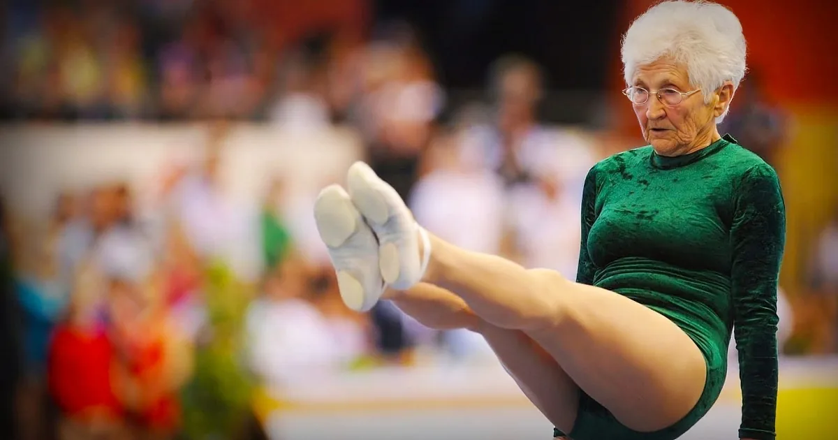 An elderly woman in a green leotard performs a gymnastics routine with her legs extended.