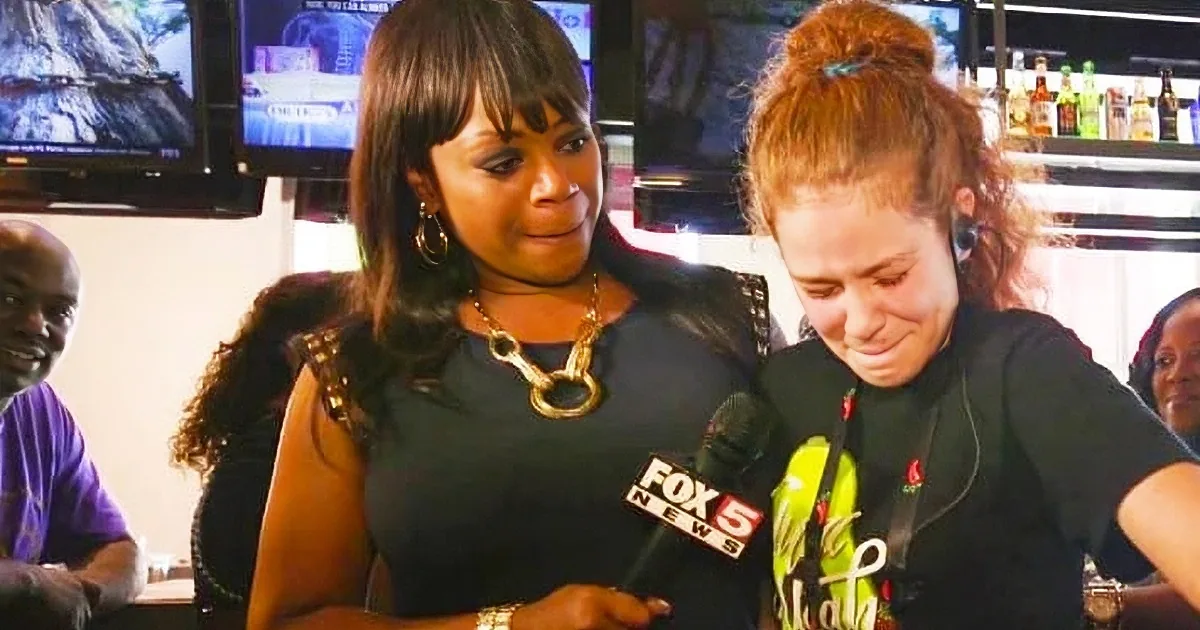 Two women are interacting closely, one holding a microphone, in a setting with other people and a bar in the background.