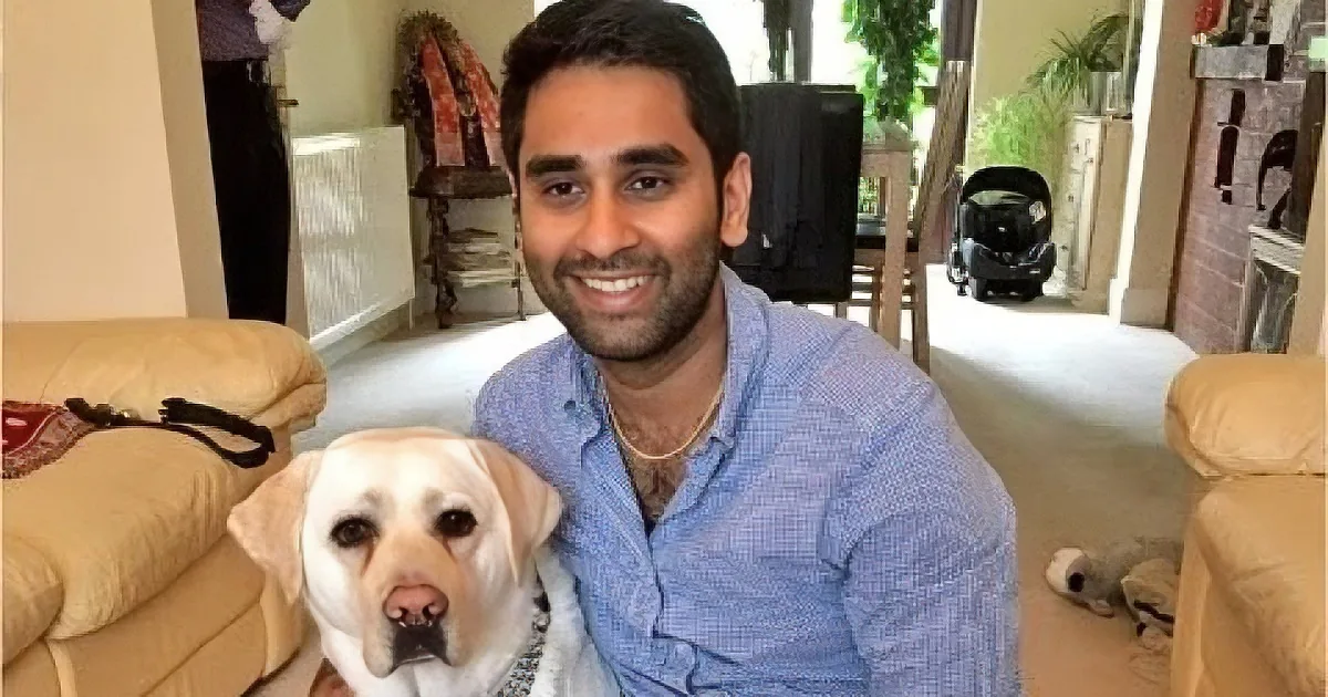 A man in a blue shirt smiles while holding a light-colored dog in a cozy living room.