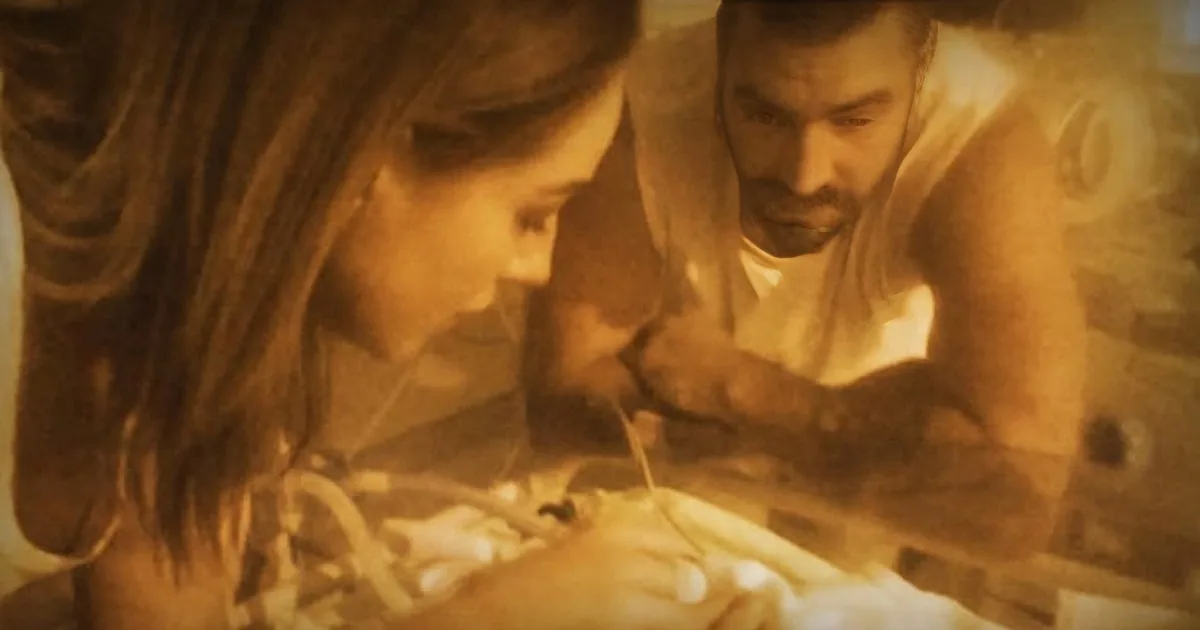 A couple leans over a newborn baby, both looking down with affection in a warm, sepia-toned setting.