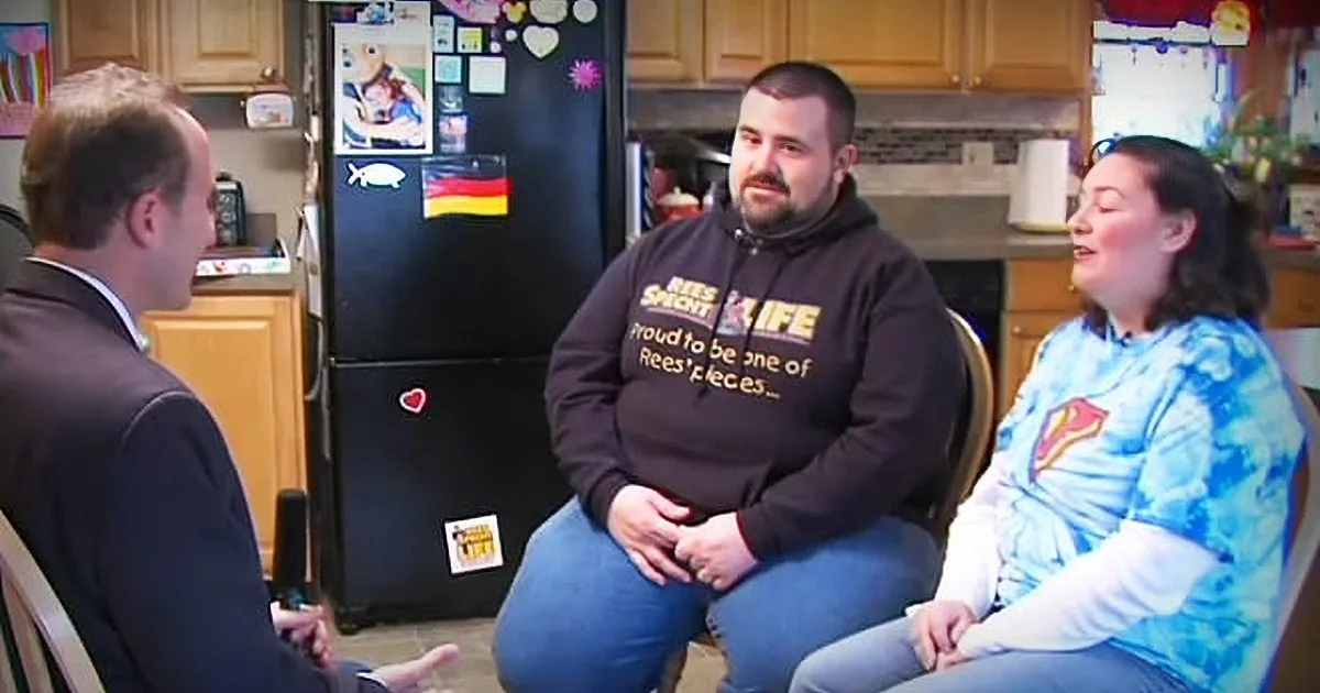 A man in a "Reese's Pieces" hoodie sits between a woman in a blue tie-dye shirt and a man in a suit in a kitchen.