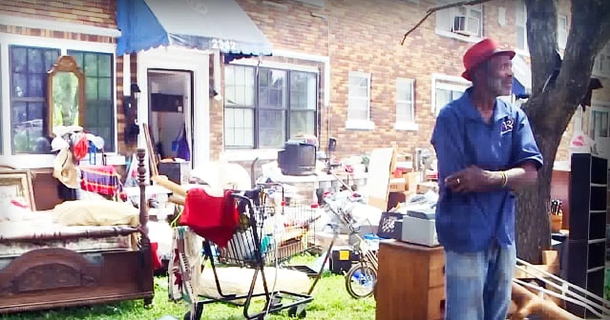 A man in a red hat stands amidst a cluttered yard sale, surrounded by various items and furniture.