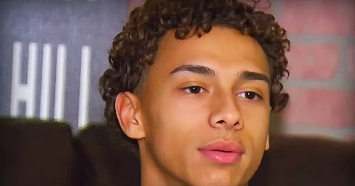 A person with curly hair sits in front of a brick wall, wearing a dark shirt.