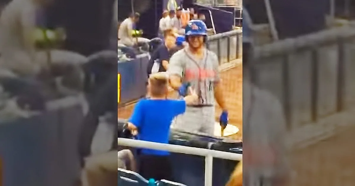 A man in a blue helmet and gray shirt is holding a red object, possibly a bat, in a stadium setting.
