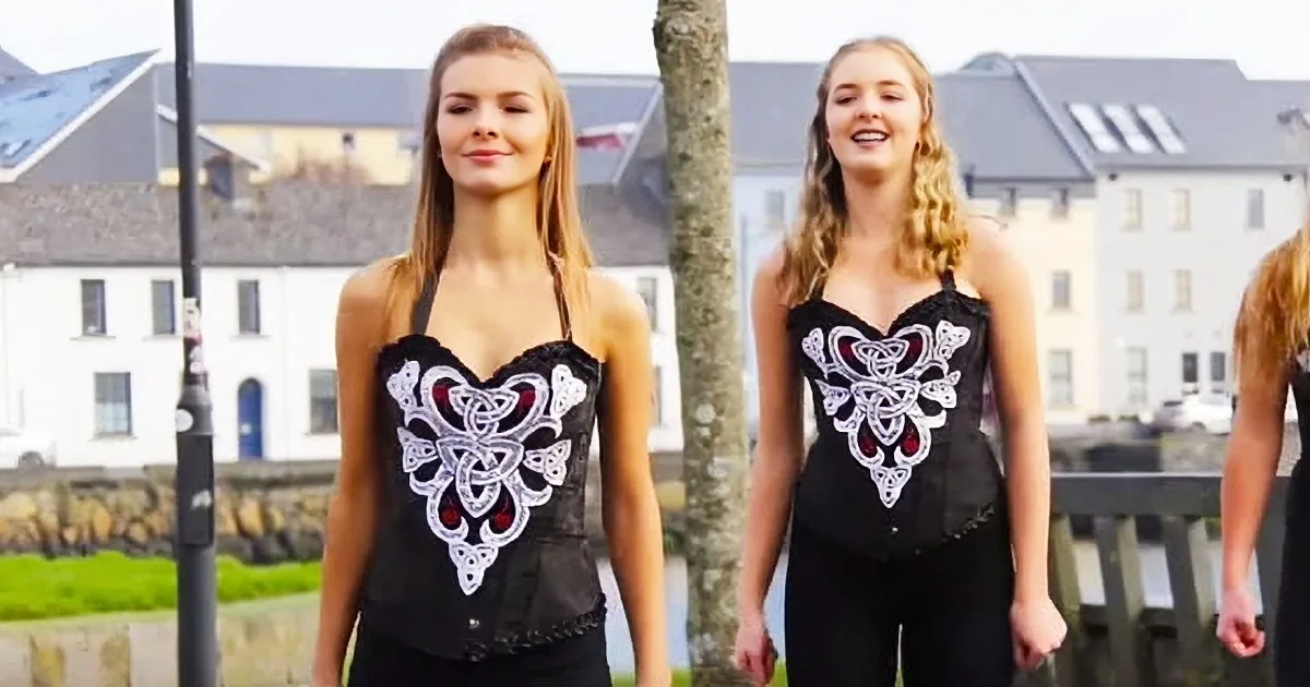 Two women in matching black outfits with white and red designs stand outdoors with buildings and trees in the background.