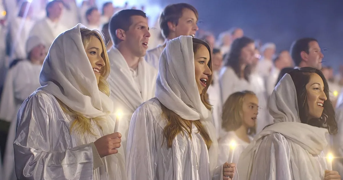 A group of people in white robes and head coverings hold candles, singing in a lit setting.