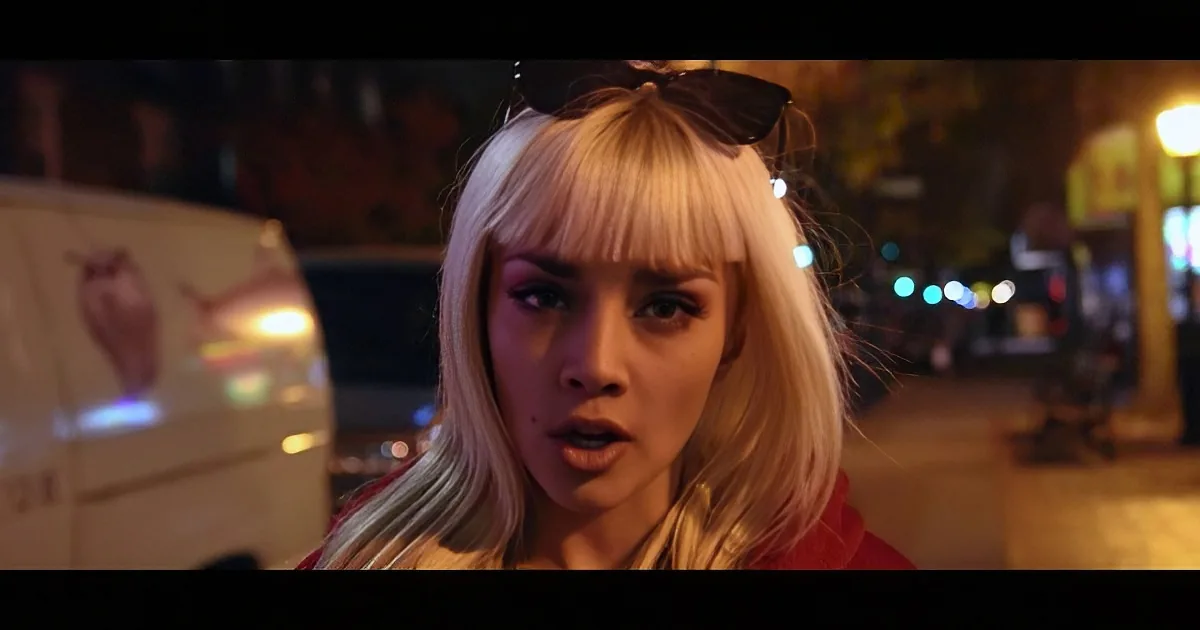 A person with blonde hair and bangs, wearing a red top, stands on a street at night with a van and blurred lights in the...