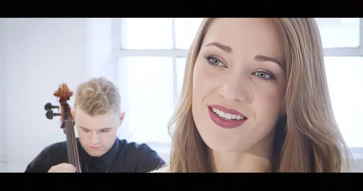 A woman smiles in the foreground while a man plays a cello in the background.