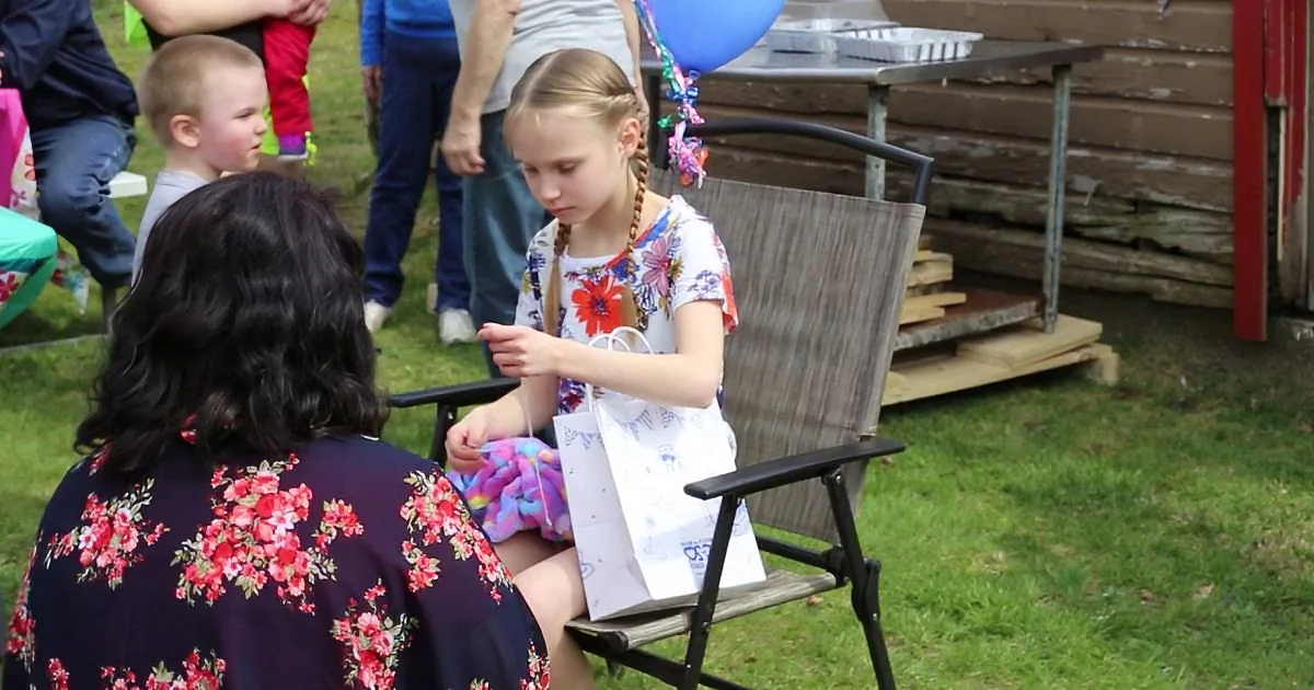 A young girl in a floral dress sits on a chair, holding a gift bag and a string, with a woman in a floral dress beside her.
