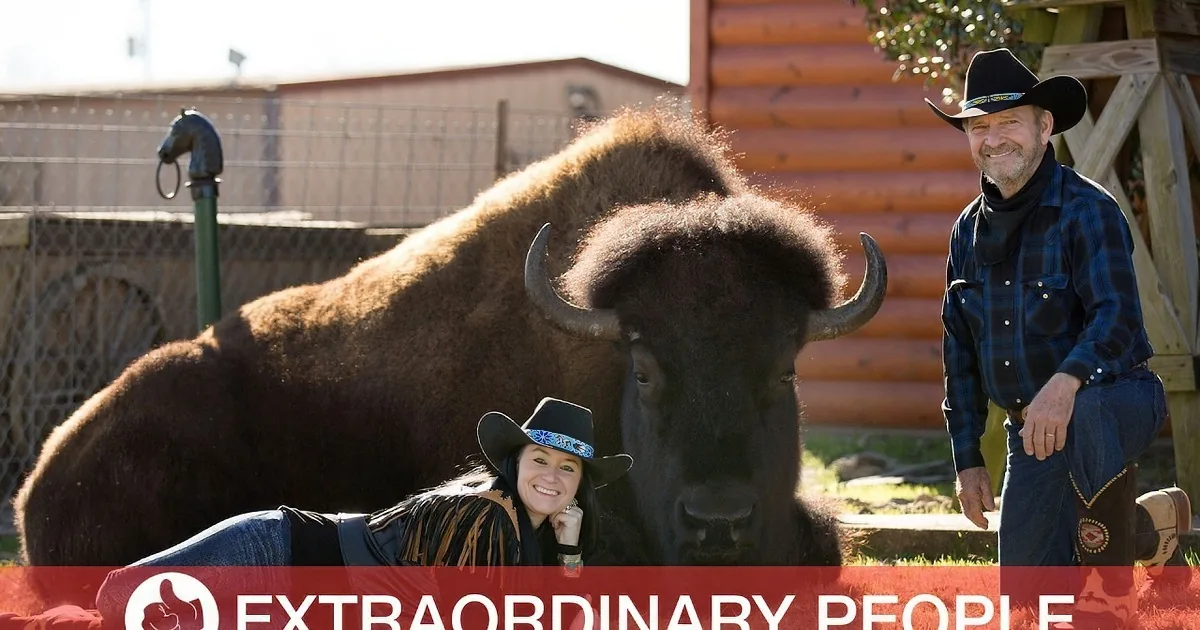 A man and woman in cowboy attire pose with a bison in a rustic setting.