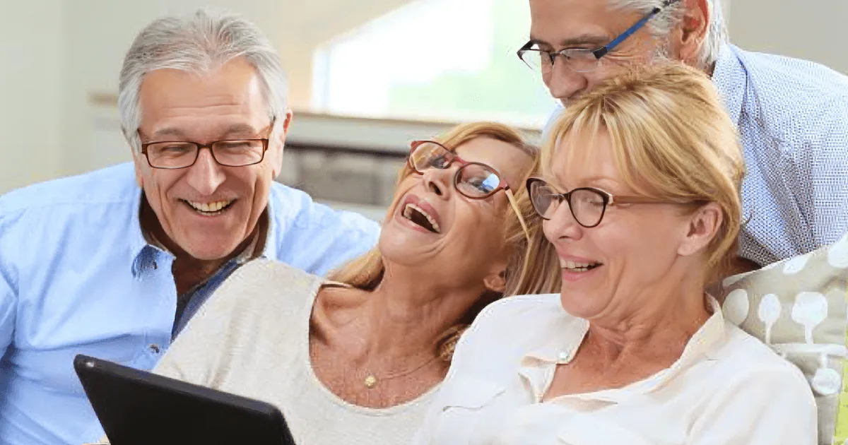 Four older adults are laughing while looking at a tablet together.