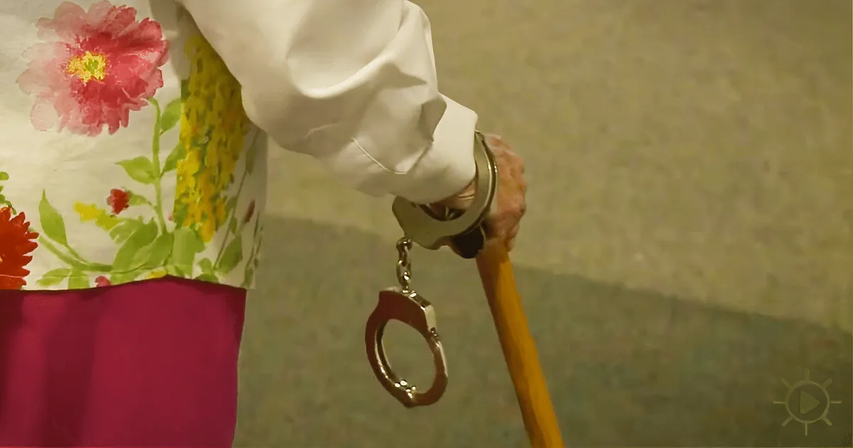A person in a floral shirt and pink skirt holds a cane with handcuffs attached.