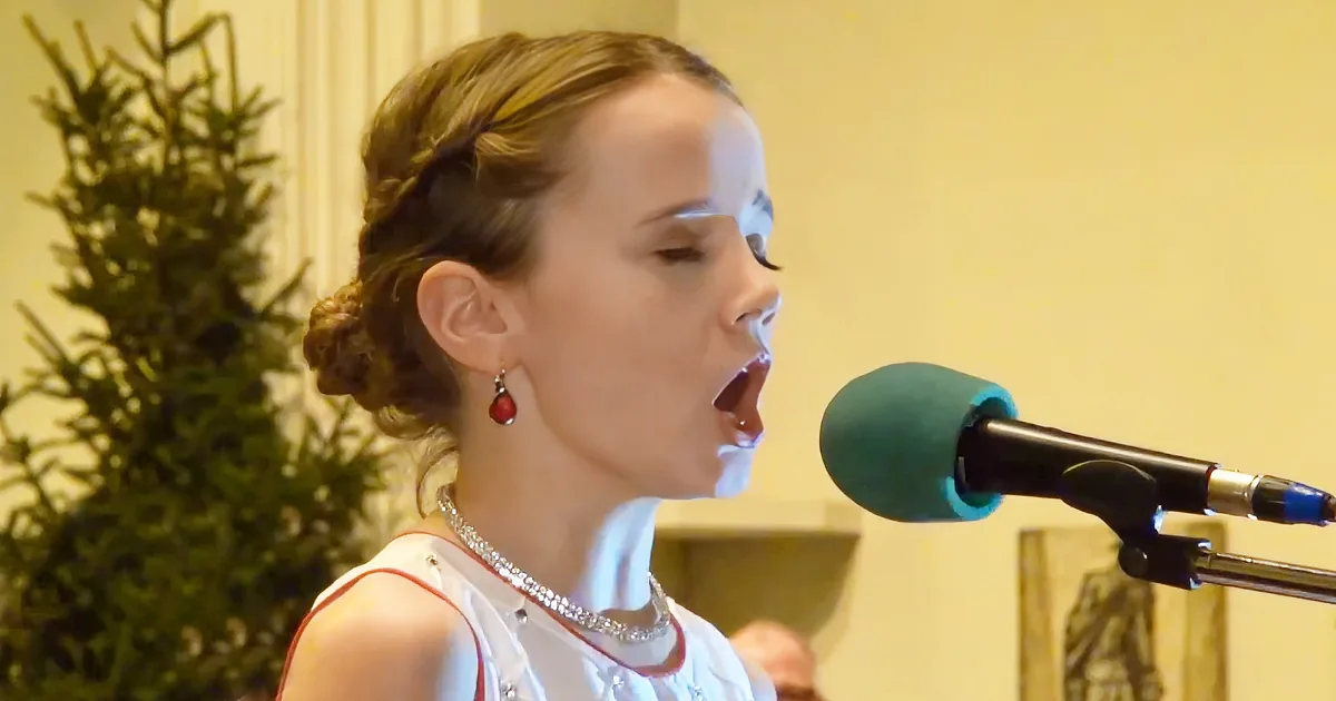 A young girl with braided hair and red earrings sings into a microphone, wearing a white outfit with a sparkly necklace.