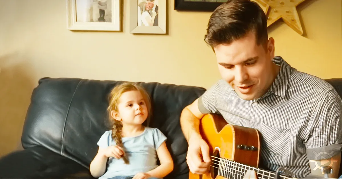 A man plays guitar while a young girl watches attentively on a black leather couch.