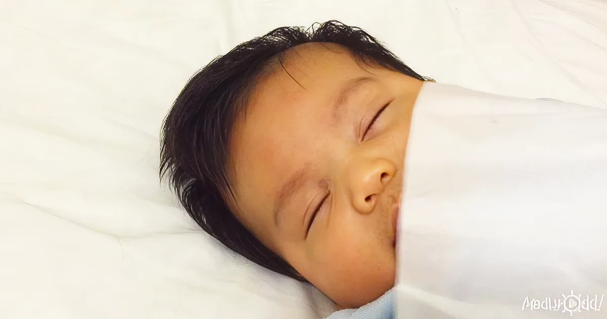 A baby with dark hair is peacefully sleeping on a white surface.