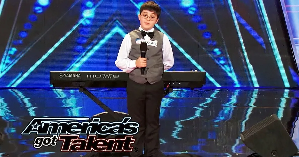 A young boy in a suit stands on stage with a microphone, preparing to perform on America's Got Talent.