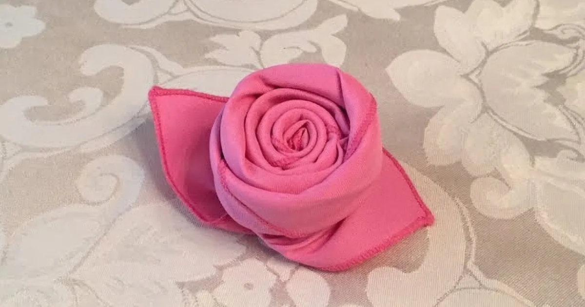 A vibrant pink fabric rose with a purple center is displayed on a light, patterned fabric background.