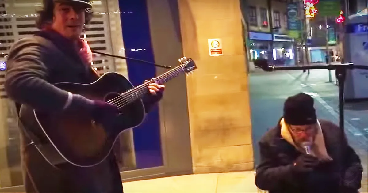 A man plays guitar on a city street while another man sits nearby, bundled up.