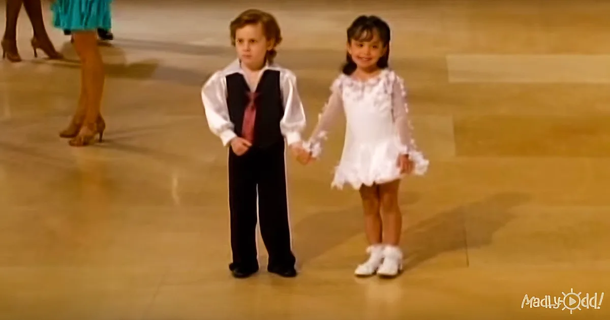 A young boy and girl hold hands, dressed formally, standing on a dance floor.