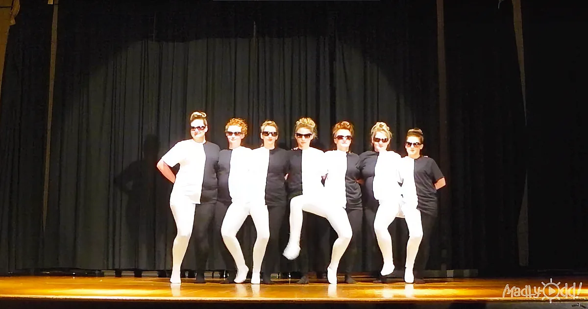 A group of dancers in white and black outfits perform on stage, wearing sunglasses.