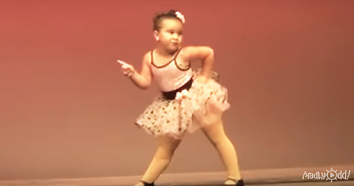 A young girl in a white dress and black tank top is dancing on stage, pointing to the left.