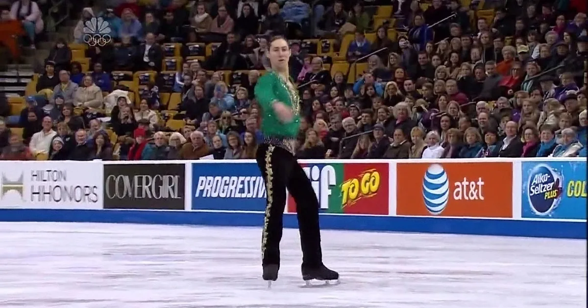 A figure skater in a green and black outfit performs a spin on the ice in front of a large audience.