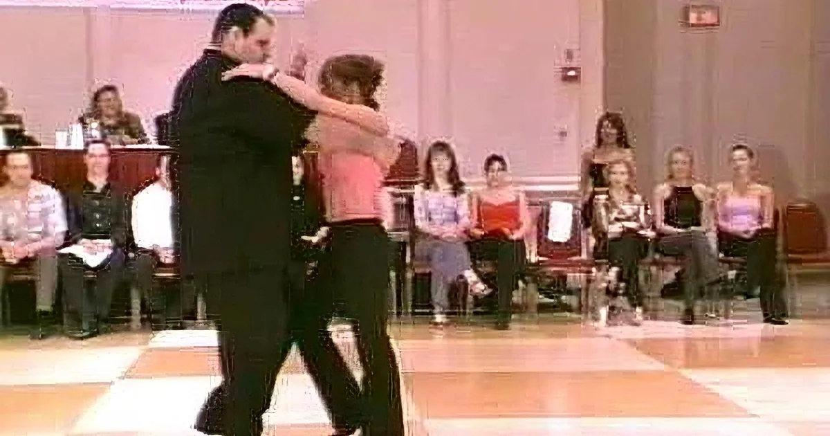 A couple in formal attire dances gracefully on a checkered floor, surrounded by spectators.