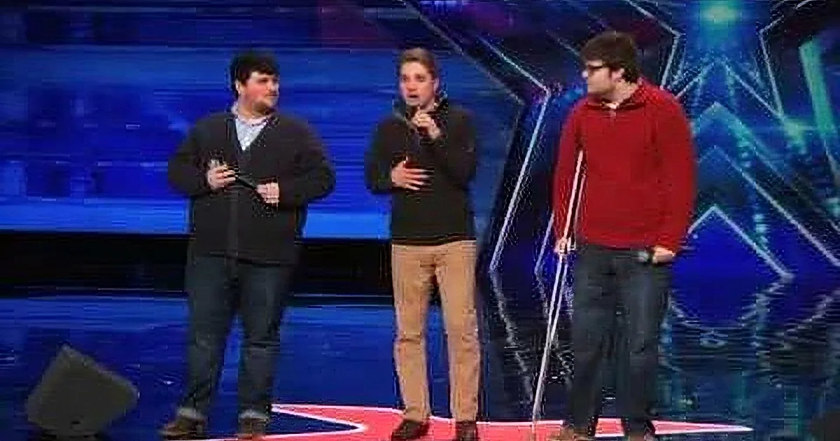 Three men in a stage setting, one in a red shirt, another in a black sweater, and the third in a black shirt, appear to...