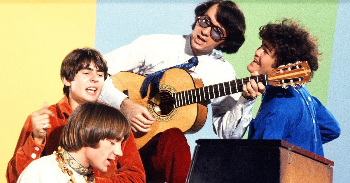 A man in a white shirt and sunglasses plays guitar while three others sing and play instruments.