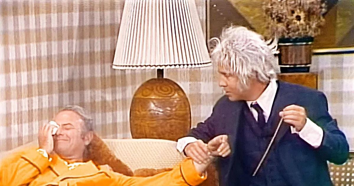 A man in a suit is sitting on a couch, holding a microphone, while another man in a robe lies on the couch, appearing to...