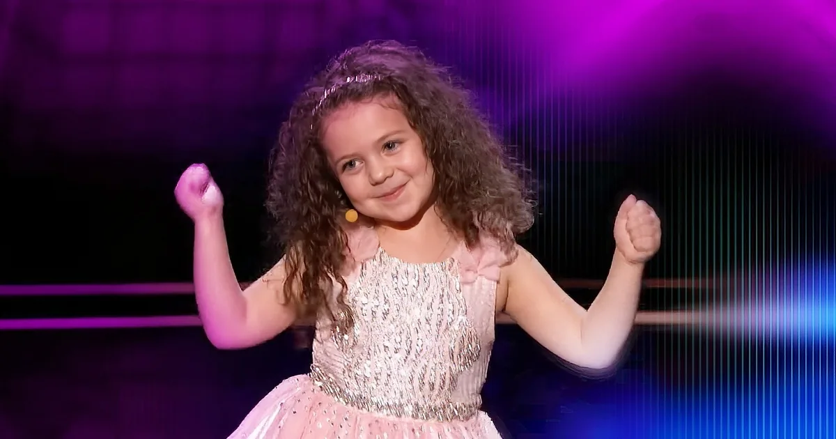 A young girl with curly hair, wearing a sparkly pink dress, smiles and raises her fists in a confident pose on stage.