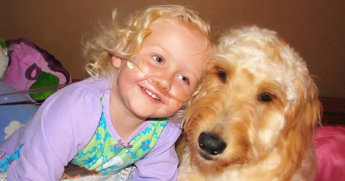 A young girl with curly blonde hair smiles next to a fluffy golden dog, both appearing happy and close.