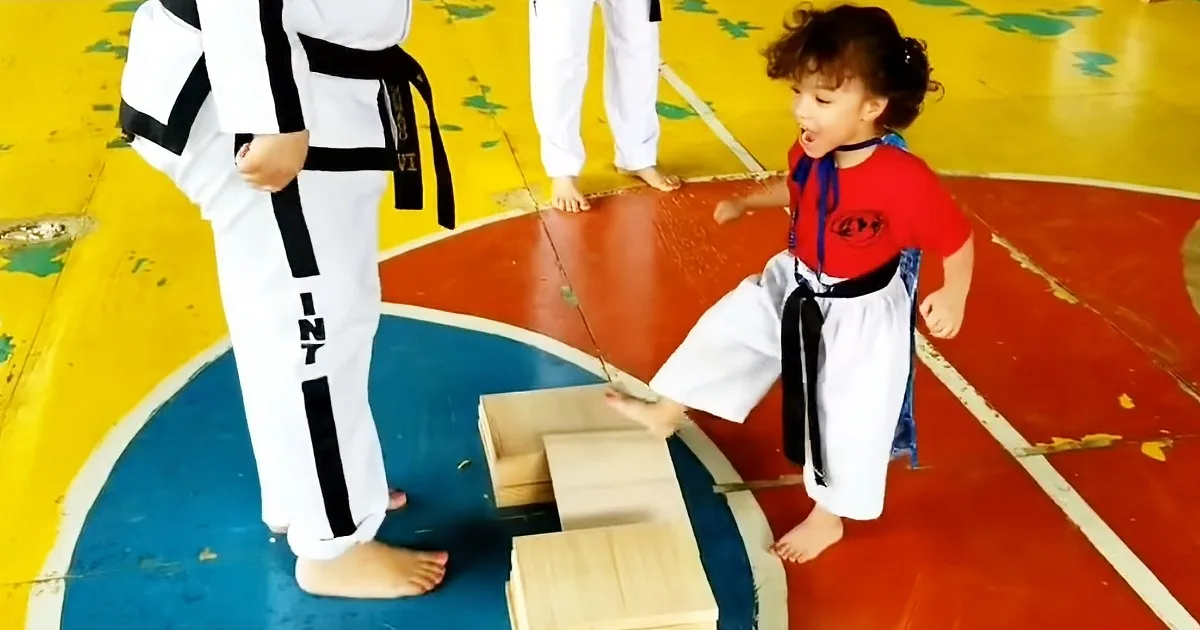 A young child in a red shirt and white pants practices a kick on a wooden board with an adult in a white uniform standing...