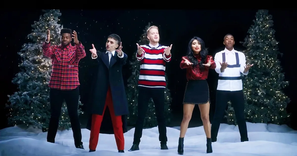 A group of five people in festive attire stands in a snowy scene with Christmas trees behind them.
