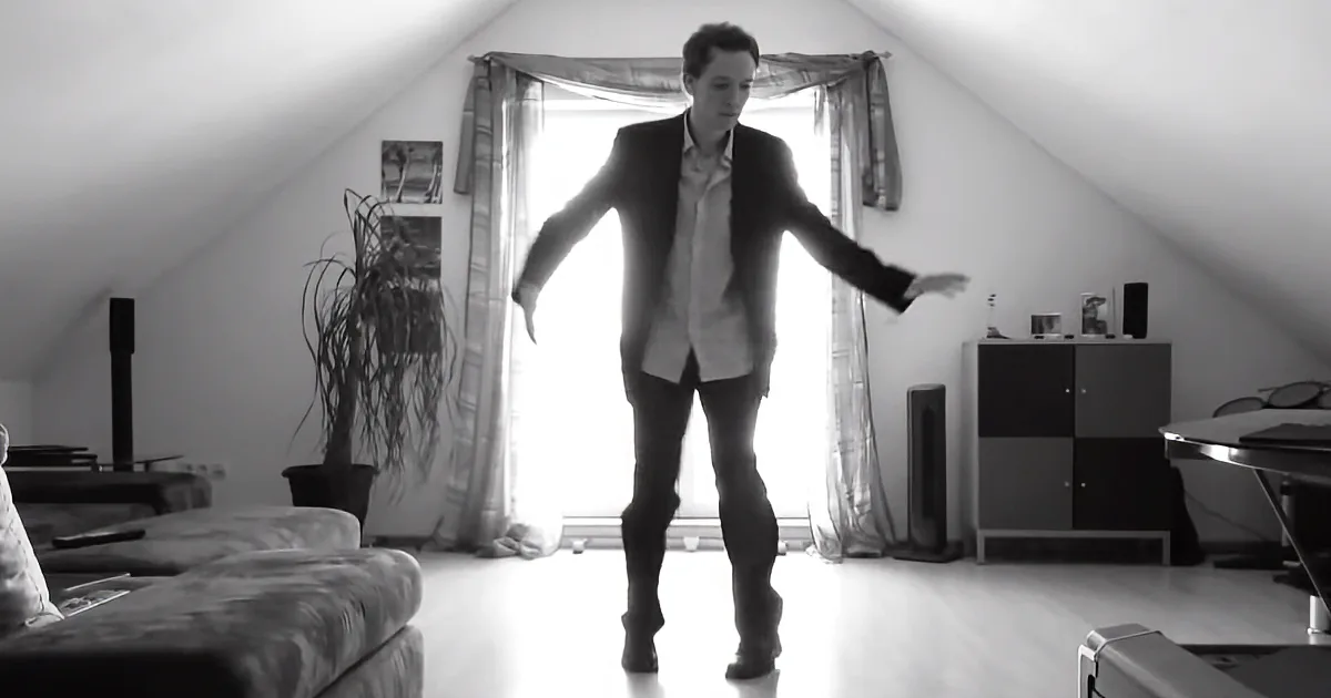 A man in a suit stands in a room with an angled ceiling, arms outstretched, near a window with curtains.