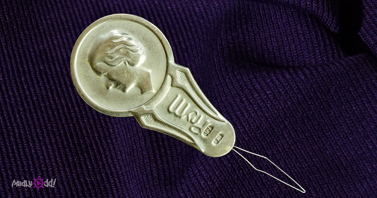 A metallic spoon-shaped object with a face and "BREM" text is placed on a purple knitted fabric.