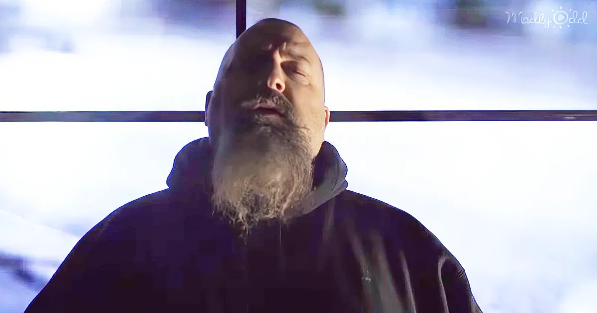 A man with a white beard and mustache, wearing a dark hoodie, stands against a bright, cloudy sky.