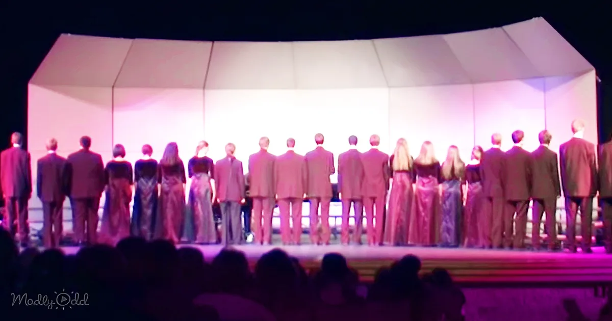 A choir stands in a line on a stage, facing away from the audience, with soft lighting creating a serene atmosphere.