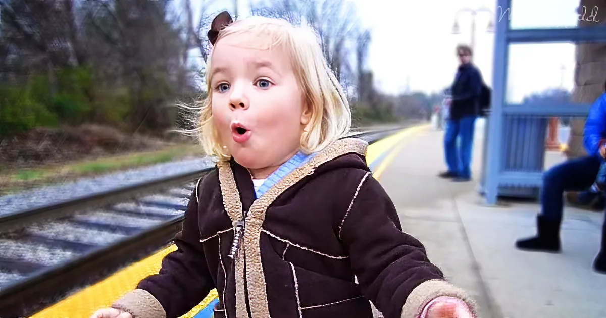 A young child with blonde hair stands on a train platform, wearing a brown jacket, with a surprised expression.