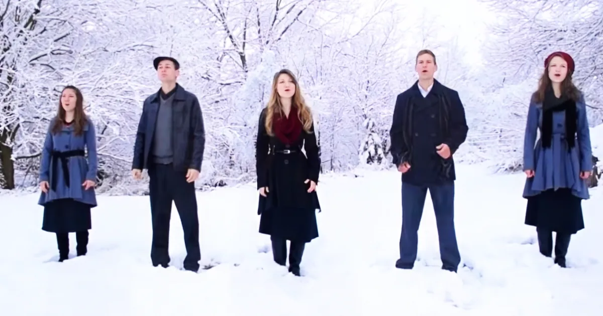 Five people in winter coats stand in a snowy forest, singing.