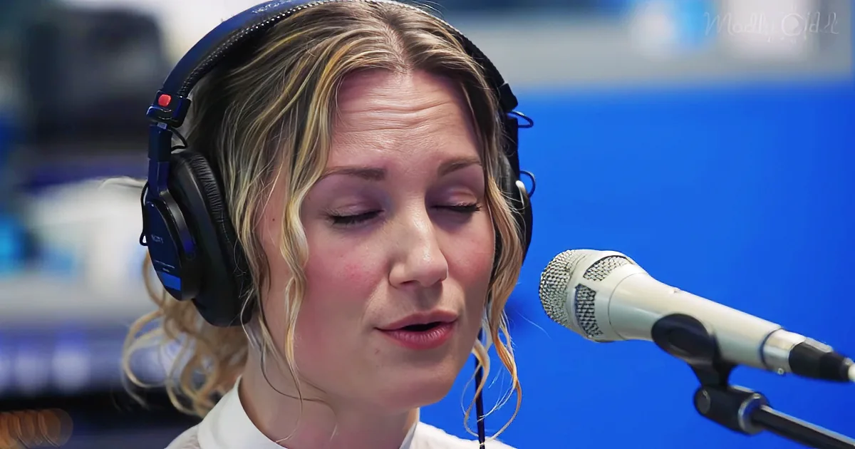 A woman with blonde hair in a ponytail, wearing headphones, sings into a microphone in a studio setting.