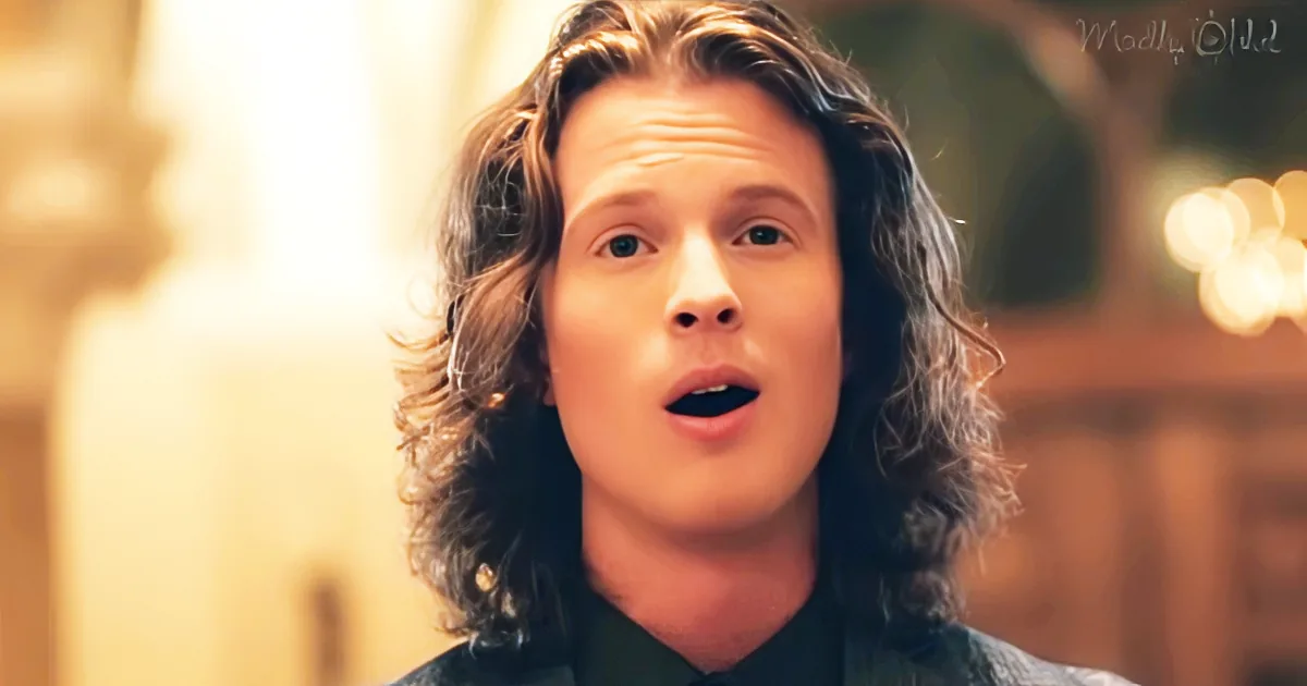 A person with long, wavy hair and a black shirt, mouth slightly open, in a warmly lit setting.