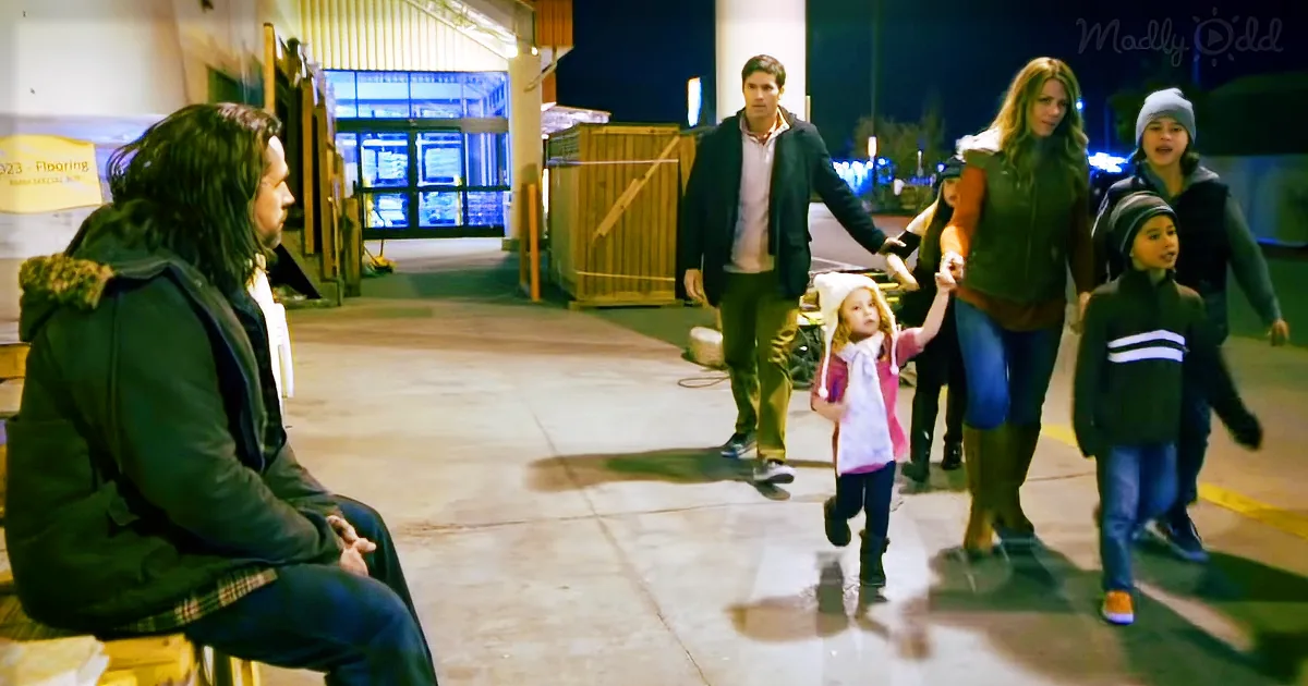 A man sits on a bench, watching a family of five walk away in a parking lot at night.