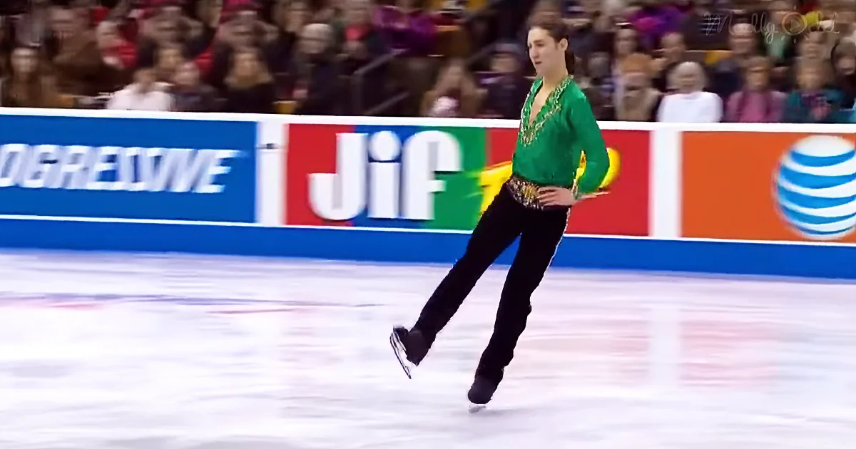 A figure skater in a green top and black pants glides on ice, with a colorful advertisement backdrop and spectators in...