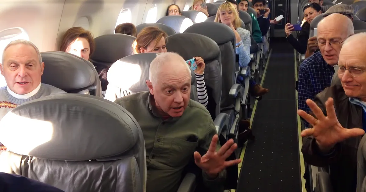 A man in a green shirt gestures animatedly on an airplane, surrounded by passengers.