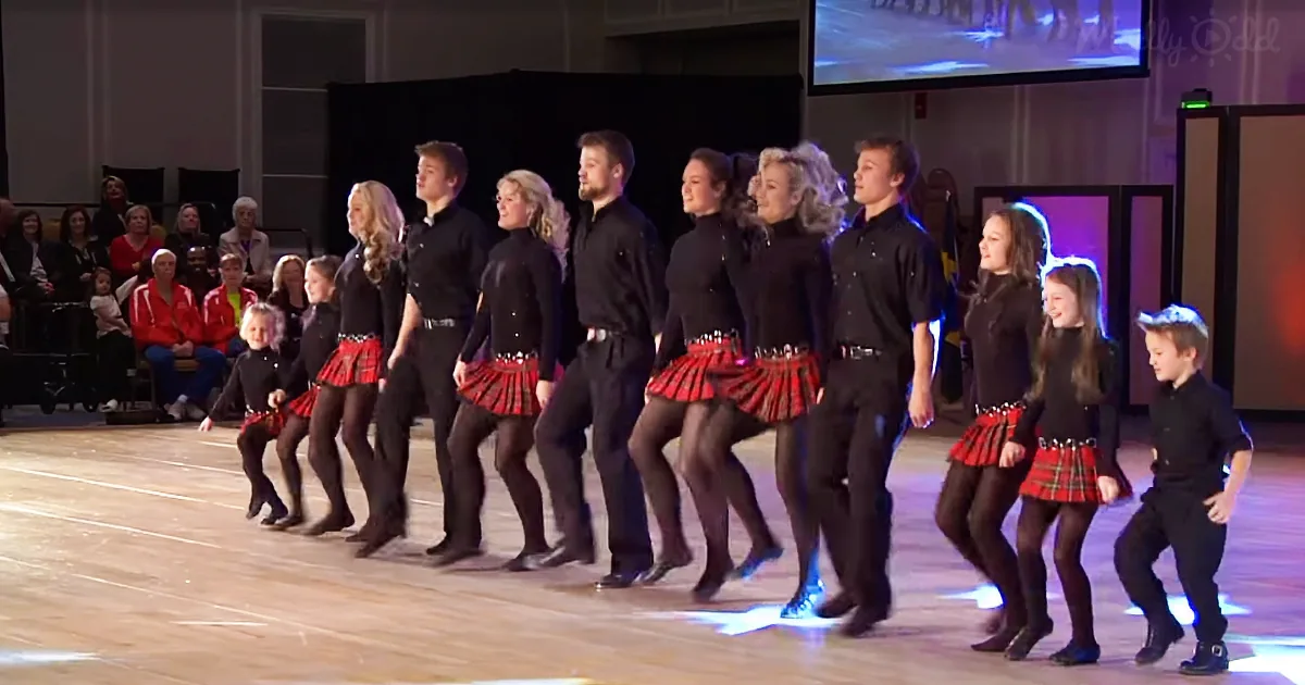 A group of dancers in black and red kilts perform a synchronized routine on stage.