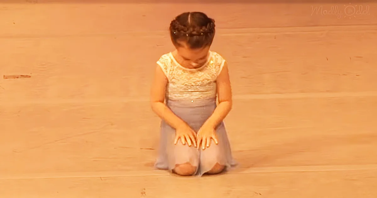 A young girl in a light-colored dress kneels on a wooden floor, hands clasped in front of her.