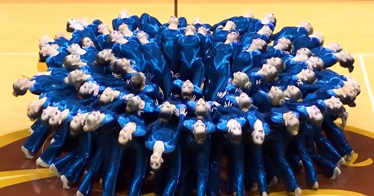 A synchronized group of dancers in blue costumes perform a coordinated pose on a basketball court.