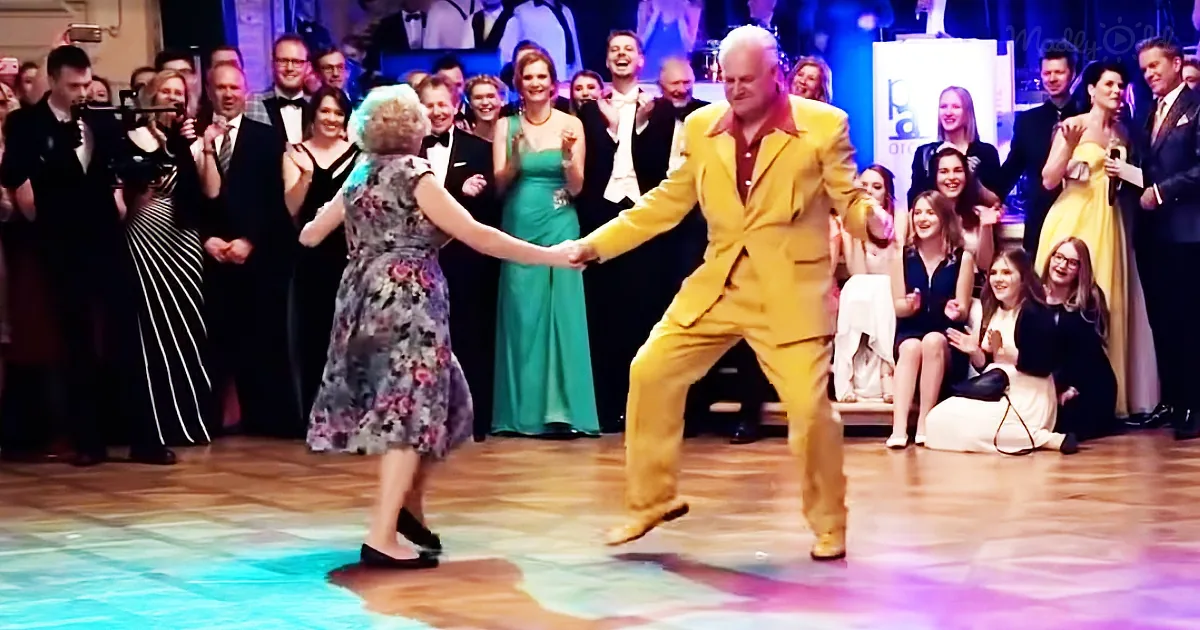 A man in a yellow suit dances with a woman in a floral dress on a stage, surrounded by a cheering crowd.